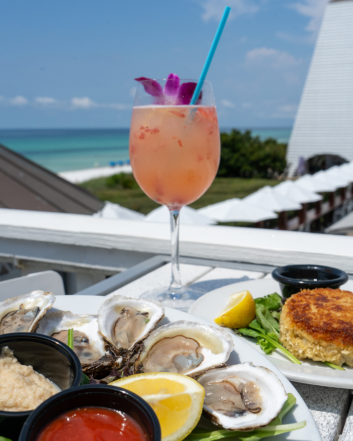 A plate of oysters and a drink with a purple flower on a table overlooking the beach