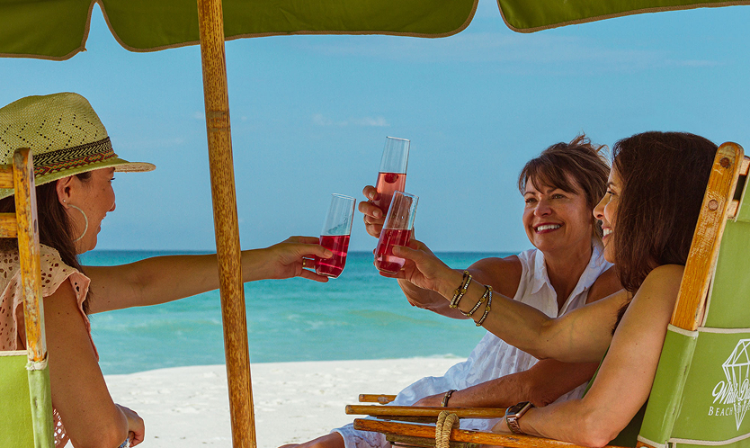 Three women drinking wine in beach chairs under umbrella